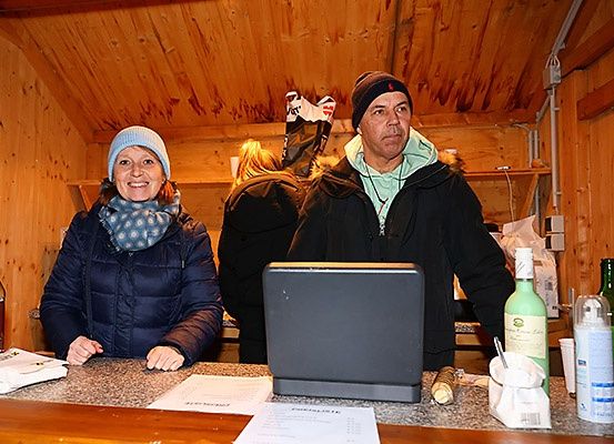 Zwei Personen stehen lächelnd hinter einer Theke. Ein Laptop, Flaschen und ein Becher stehen auf der Theke. Eine Frau trägt eine blaue Jacke und eine Mütze, während ein Mann eine schwarze Jacke trägt. Sie befinden sich in einem Raum mit Holzwänden.