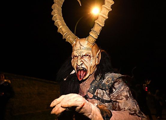 Eine Person in einer gehörnten Maske und einem langhaarigen Kostüm erscheint bedrohlich, mit offenem Mund und herausgestreckter Zunge, beleuchtet von einem Licht.