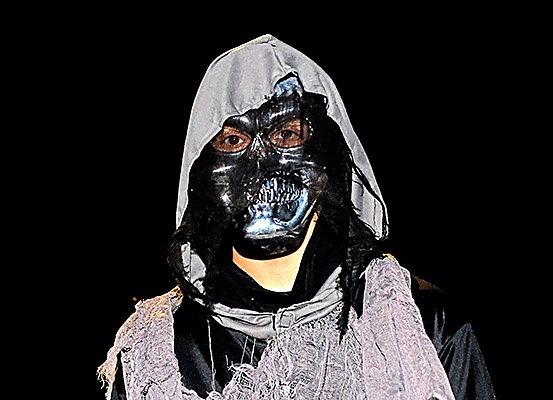 Eine Person in einem schwarzen und grauen Kostüm mit einer schädelartigen Maske vor dunklem Hintergrund.