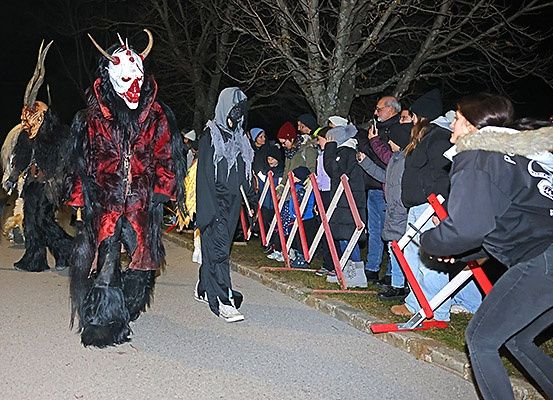 In der Nacht gehen Menschen in Kostümen auf der Straße. Zuschauer beobachten sie und einige machen Fotos. Rote Barrieren sind am Straßenrand aufgestellt.