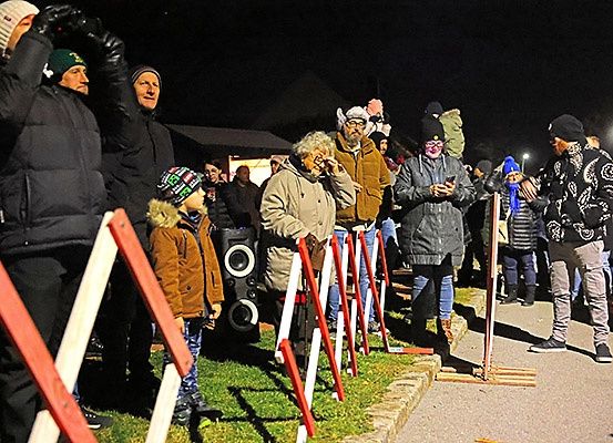 Eine Gruppe von Menschen steht bei Nacht in einer Reihe, einige tragen Mützen und Handschuhe. Sie scheinen eine Veranstaltung zu beobachten. Das Gebiet ist von roten und weißen Barrieren umgeben.