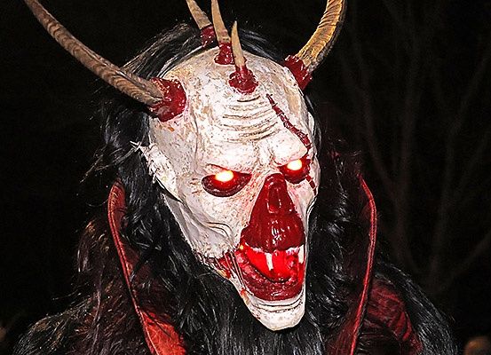 Eine Person in einem roten Umhang, die eine weiße hörnige Maske mit roten Augen und offenem Mund trägt, posiert im Dunkeln.