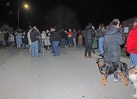Eine Menschenmenge versammelt sich nachts in einer Schlange am Straßenrand. Einige von ihnen tragen Mützen und Turnschuhe. Ein Hund steht am Straßenrand.