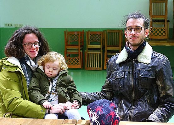 Eine Familie mit drei Personen sitzt in einem Raum mit grünen Wänden, eine Frau mit Brille, ein Kind mit geschlossenen Augen und ein Mann in einer schwarzen Jacke mit weißem Pelzkragen. Im Hintergrund stehen Holzstühle und ein Holztisch.