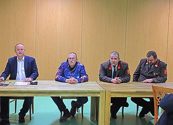 Vier Männer sitzen an einem Tisch, einer trägt eine Brille und ein anderer ein Militäruniform. Sie blicken nach vorne, möglicherweise in einer Besprechung oder Konferenz.