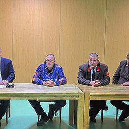 Vier Männer sitzen an einem Tisch, einer trägt eine Brille und ein anderer ein Militäruniform. Sie blicken nach vorne, möglicherweise in einer Besprechung oder Konferenz.