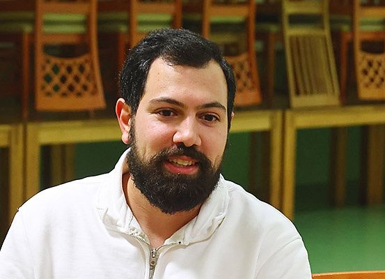 Ein Mann mit Bart sitzt lächelnd in einem Klassenzimmer, mit Holzstühlen und einer grünen Wand im Hintergrund.