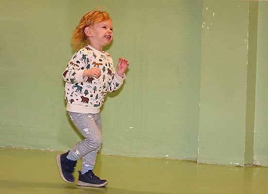 Ein junges Kind mit roten Haaren tanzt in einem Raum mit grünen Wänden und einem gelben Boden. Das Kind trägt ein weißes Langarmshirt mit Tierdruck und graue Hosen.
