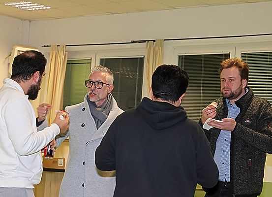 Vier Männer stehen in einem Raum, möglicherweise in einer Besprechung. Sie halten Essen und scheinen miteinander zu sprechen. Der Raum hat Glasfenster mit Vorhängen.
