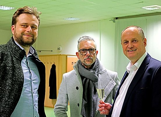 Drei Männer stehen in einem Raum zusammen. Der Mann auf der linken Seite trägt einen schwarzen Mantel. Der Mann in der Mitte trägt eine Brille und hält ein Champagnerglas. Der Mann auf der rechten Seite trägt einen dunklen Anzug.