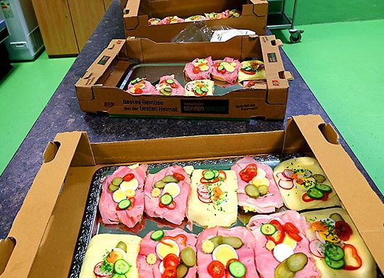 Mehrere offene Kartons auf einer Arbeitsplatte, gefüllt mit Sandwiches, die mit verschiedenen Gemüsesorten und Fleisch, möglicherweise Schinken, belegt sind.
