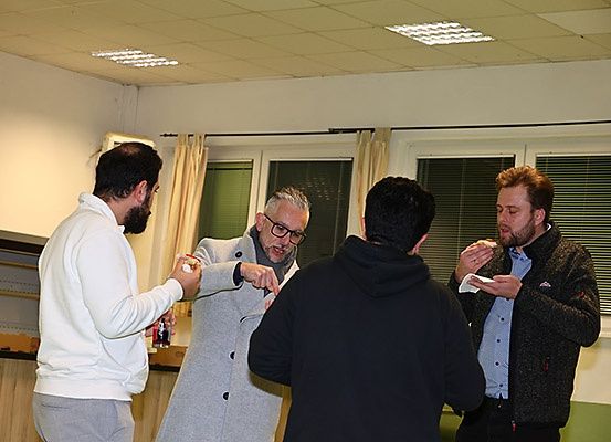 Vier Männer sind drinnen versammelt, möglicherweise in einer Besprechung. Der Mann auf der linken Seite hält ein kleines Objekt, möglicherweise Essen, und gestikuliert mit der anderen Hand. Der Mann in der Mitte trägt eine Brille und schaut den Mann auf der rechten Seite an. Der Mann auf der rechten Seite hält ein Stück Papier und scheint zu sprechen.