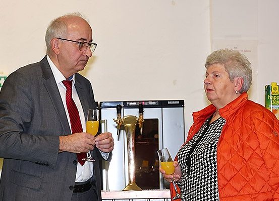 Ein älterer Mann und eine Frau stoßen mit orangefarbenen Getränken in einem Raum mit einer Bareinrichtung und einer weißen Wand an.