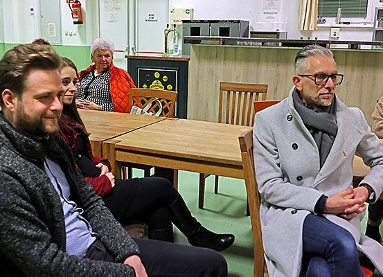 Eine Gruppe von Menschen sitzt um einen Tisch in einem Raum, wahrscheinlich einem Café. Ein Mann mit Brille und Schal sitzt vorne, während andere dahinter sitzen. Es gibt Holzstühle und eine Theke mit einem Feuerlöscher im Hintergrund.