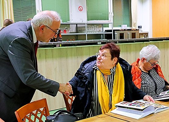 Ein Mann im Anzug schüttelt die Hand einer Frau in einem gestreiften Mantel. Sie hält ein Buch. Hinter ihnen sitzen zwei andere Frauen an einem Tisch, eine blickt in ein Buch. Im Hintergrund ein Tresen und ein Schild mit der Aufschrift 'Nein'.
