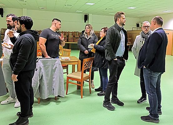 Eine Gruppe von Menschen steht in einem Raum, wahrscheinlich bei einer Besprechung. Einige halten Tassen und Teller, während andere in Gespräche vertieft sind. Der Raum hat Holzstühle, einen Schreibtisch und einen grünen Boden.