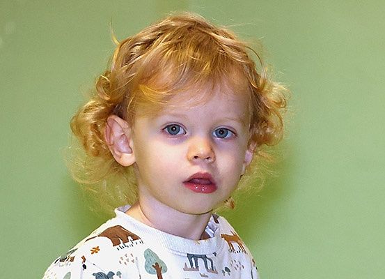 Ein junges Kind mit lockigem blondem Haar und blauen Augen schaut direkt in die Kamera und trägt ein weißes Shirt mit Tieraufdrucken.