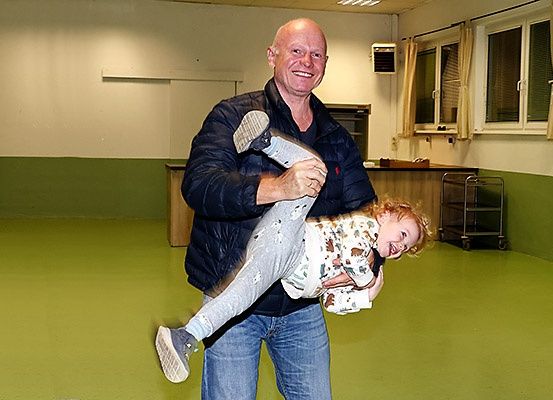 Ein Mann hält ein lächelndes Kind in einem grünen Raum mit grünem Boden, weißen Wänden und zwei Fenstern.