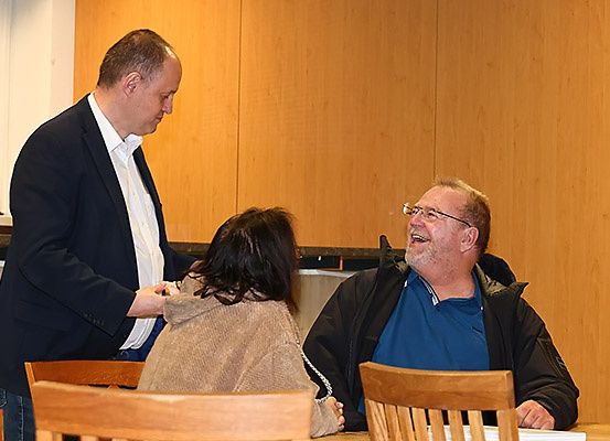 Drei Personen sitzen an einem Tisch, zwei lächeln. Der Mann in formeller Kleidung steht hinter der Frau. Der Mann mit Brille lacht. Holzstühle und eine Holzwand sind im Hintergrund.
