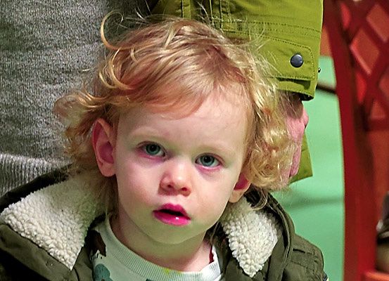 Ein blondes Kind mit blauen Augen schaut mit rosa Lippen nach vorne. Hinter dem Kind trägt eine Person eine grüne Jacke.