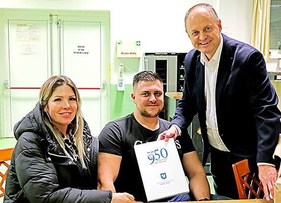 Drei Menschen lächeln für ein Foto, einer hält ein Zertifikat. Der Mann hat einen muskulösen Körperbau. Dahinter steht ein Feuerlöscher und rechts steht ein Kühlschrank.
