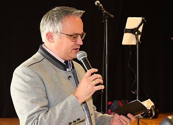 Ein älterer Mann mit Brille spricht in ein Mikrofon und hält ein Buch. Ein Mikrofonständer steht hinter ihm.