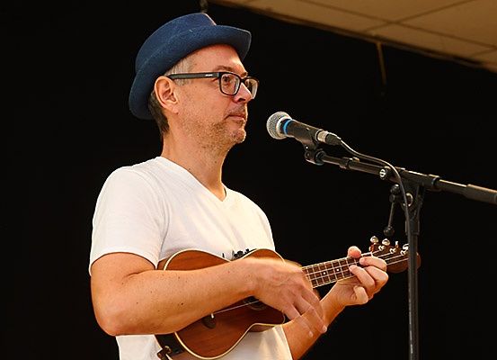 Ein Mann mit Brille und Hut spielt eine Ukulele und singt in ein Mikrofon auf der Bühne.