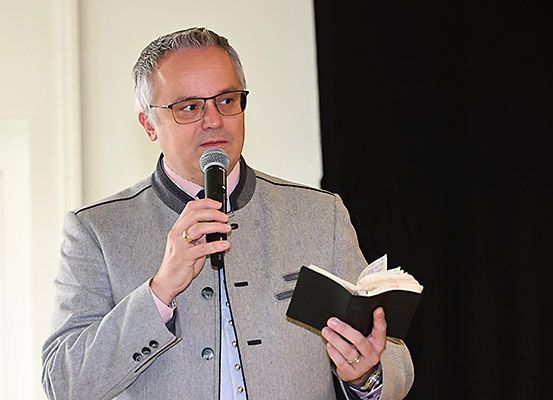 Ein Mann in einem grauen Anzug spricht in ein Mikrofon und hält ein Buch in der Hand, vermutlich während eines Gottesdienstes.