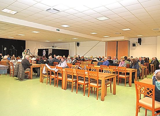 Ein Innenbereich mit vielen Menschen, die auf Stühlen um Tische herum sitzen. Einige Personen stehen und unterhalten sich. Es gibt Lautsprecher und eine Bühne mit einem Vorhang.
