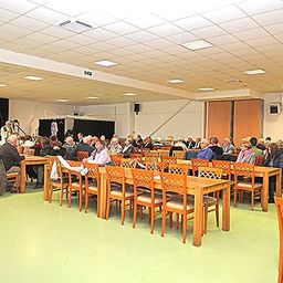 Ein Innenbereich mit vielen Menschen, die auf Stühlen um Tische herum sitzen. Einige Personen stehen und unterhalten sich. Es gibt Lautsprecher und eine Bühne mit einem Vorhang.