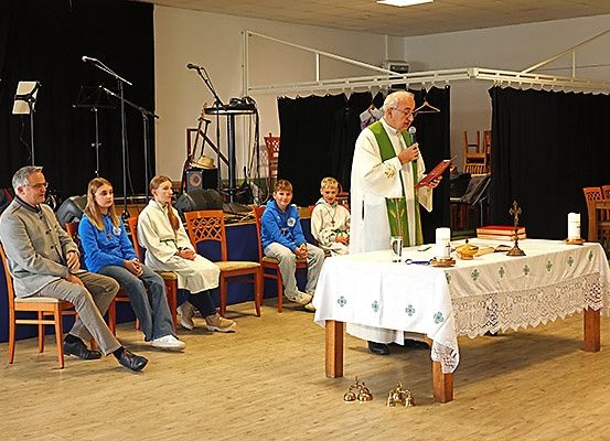 Ein Priester steht an einem Altar und spricht in ein Mikrofon. Menschen sitzen auf Stühlen vor ihm und lauschen.