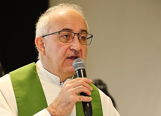 Ein älterer Mann in einer grünen und weißen Kirchenkleidung spricht in ein Mikrofon. Er hält das Mikrofon mit der rechten Hand.