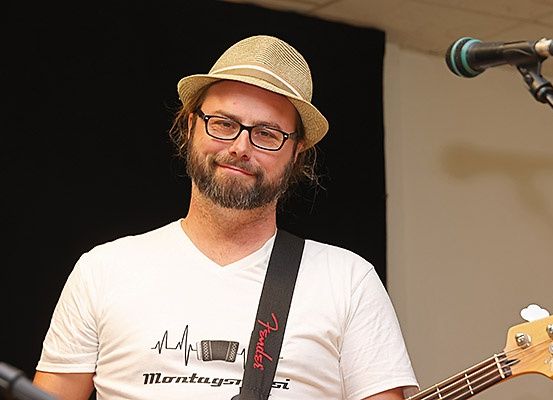 Ein Mann mit Bart, der einen Hut und eine Brille trägt, spielt eine Bassgitarre vor einem Mikrofon.