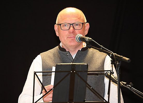 Ein Mann in einer Brille und einer Weste steht vor einem Mikrofon auf einem Ständer und liest von einem Stück Papier.
