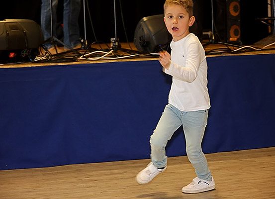 Ein junger Junge tritt auf der Bühne auf, trägt ein weißes Shirt und hellblaue Jeans. Er hält ein kleines Objekt in der Hand.