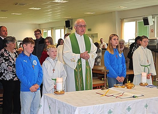 Ein älterer Priester in grünen und weißen Gewändern leitet eine Gebetszeremonie mit mehreren Kindern und Erwachsenen in einer Kirche.
