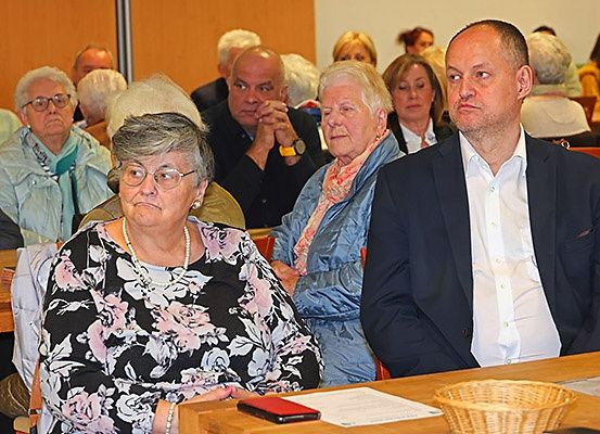 Eine Gruppe älterer Menschen sitzt in einem Konferenzraum. Eine Frau trägt eine Brille und eine Halskette. Eine andere Frau trägt einen Schal. Ein Mann im Anzug sitzt neben ihr.