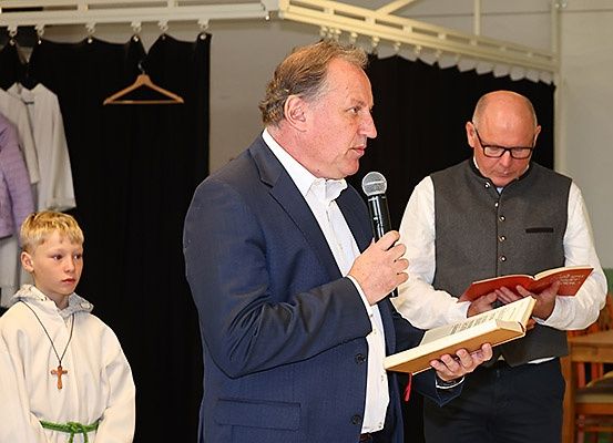 Ein Mann in einem blauen Anzug spricht in ein Mikrofon, während ein anderer Mann in einem weißen Hemd und einer Weste ein Buch liest. Ein junger Junge in einem weißen Gewand und einem Kreuzanhänger steht in der Nähe.