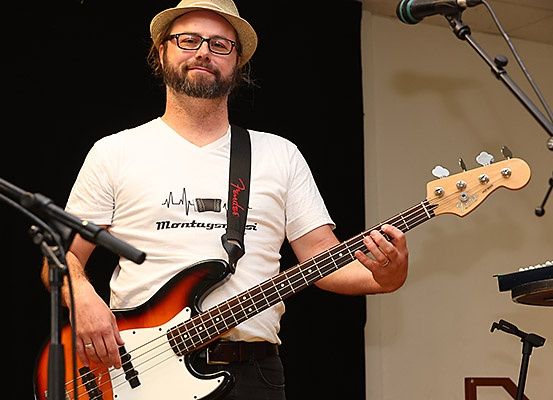 Ein Mann spielt auf der Bühne eine Bassgitarre und trägt eine Mütze und eine Brille.