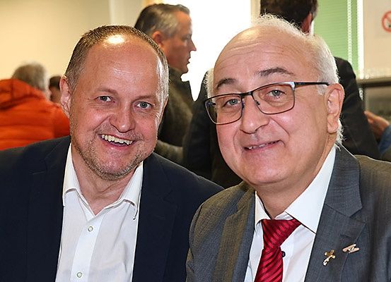 Zwei Männer in Anzügen lächeln für die Kamera mit einer Menschenmenge im Hintergrund.