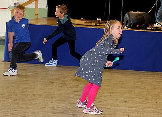 Drei Kinder laufen fröhlich auf einem Holzboden und halten grüne Objekte in den Händen. Dahinter befindet sich eine blaue Bühne mit einem Lautsprecher. Eine Person steht rechts, trägt Jeans und Sneakers.