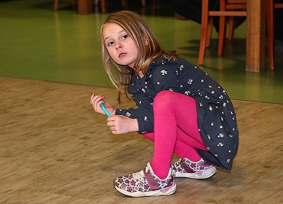Ein kleines Mädchen in einem Blumenhemd und pinken Leggings kniet auf einem Holzboden und hält einen blauen Gegenstand.