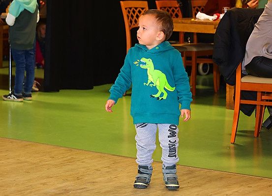 Ein Kleinkind in einer Dinosaurier-Kapuzenjacke läuft auf einem grünen Boden mit Holzstühlen und Tischen im Hintergrund.