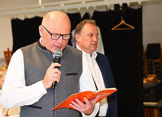 Ein Mann hält ein Mikrofon und liest ein Buch, während ein anderer Mann hinter ihm steht und das Buch ansieht. Ein Kind steht hinter ihnen.