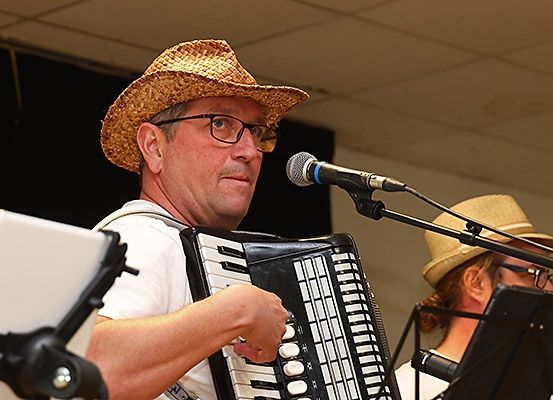 Ein Mann spielt Akkordeon auf der Bühne, trägt einen Strohhut und eine Brille, vor ihm steht ein Mikrofon. Eine weitere Person steht hinter ihm, trägt ebenfalls einen Hut und schaut nach unten.