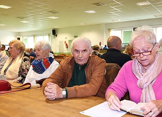 Eine Gruppe älterer Menschen sitzt um einen Tisch herum, einer liest ein Buch, während andere aufmerksam zuhören, in einem Raum mit weißen Wänden und Fenstern.