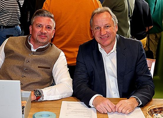 Zwei Männer sitzen lächelnd an einem Tisch, mit Papieren vor ihnen. Einer trägt eine Weste, der andere einen Anzug. Hinter ihnen stehen und plaudern Leute.