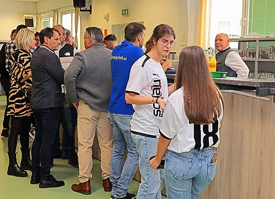 Eine Gruppe von Menschen steht in einer Reihe, wahrscheinlich bei einer Veranstaltung. Sie sind in verschiedene Kleidungsstücke gekleidet. Die Frau auf der rechten Seite trägt eine Brille und ein weißes T-Shirt mit der Nummer 18. Hinter ihnen steht ein Mann in einer Weste. Auf der Theke stehen Flaschen. Der Raum hat weiße Wände und Glasfenster.