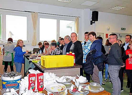 Eine Gruppe von Menschen steht in einem Raum, wahrscheinlich an einem Buffet. Einige von ihnen tragen eine Brille und andere tragen Turnschuhe. Der Tisch ist mit Tellern, Schüsseln und Tassen gefüllt, und es stehen Flaschen an der Seite. Ein gelber Aufbewahrungsbehälter steht auf der Theke, und ein Eimer steht auf dem Boden. Es gibt auch Fenster mit Vorhängen.
