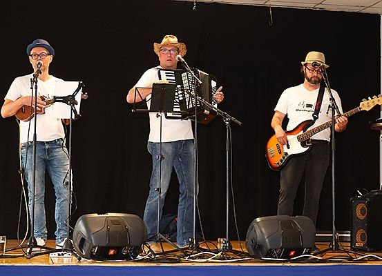 Drei Männer auf der Bühne, einer spielt Akkordeon, ein anderer Gitarre und der letzte Bassgitarre. Alle tragen Hüte und halten Mikrofone. Es gibt Lautsprecher und Musikinstrumente auf der Bühne.
